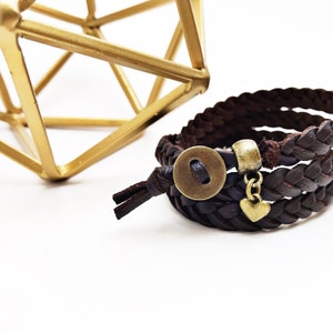 May include: Braided brown leather bracelet with a bronze-colored button clasp and a small heart charm. The bracelet is layered and has a rustic, handcrafted appearance. A gold geometric sculpture is in the background.
