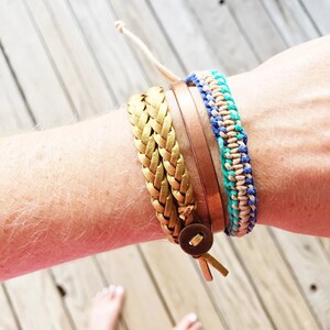 May include: A collection of bracelets in various styles. A gold braided bracelet with a wooden button closure, a thin copper-colored band, and a blue and turquoise woven bracelet are visible. A natural-colored cord is also present.