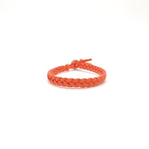 May include: A braided orange bracelet with a small clasp and a leather tie closure. The bracelet is made of woven material and is displayed on a white background. The bracelet is approximately 18 cm in circumference.