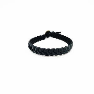 May include: A black braided leather bracelet with a brass clasp. The bracelet is made of woven leather strips, creating a textured, circular design. The bracelet is a simple, yet stylish accessory.