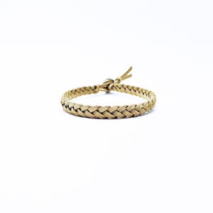 May include: Braided tan leather bracelet with a loop and knot closure. The bracelet is made of multiple strands of leather woven together, creating a textured appearance. The bracelet is shown on a white background.