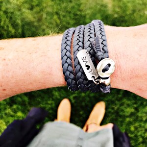 May include: Braided gray leather wrap bracelet with a silver charm that reads "LOVE" and a heart symbol. The bracelet wraps multiple times around the wrist and fastens with a silver button clasp. The bracelet is shown on a person's arm.