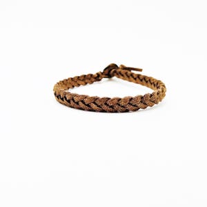 May include: Braided brown bracelet with a textured appearance. The bracelet is made of a woven material and features a knot closure. The bracelet is displayed on a white background.