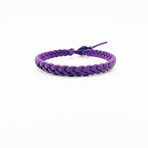 May include: A braided purple leather bracelet. The bracelet is a continuous loop, with a woven design. The leather is a solid, vibrant purple color. The bracelet is shown against a white background.