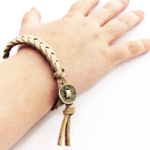 May include: A braided tan leather bracelet featuring a circular antique brass button closure. The bracelet is constructed from a single strip of leather woven into a braid, secured with a knot and a metal button that acts as a fastener.