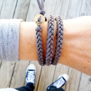 May include: Braided gray wrap bracelet with a gold-colored button closure. The bracelet is wrapped around a wrist, showcasing the woven texture and the button detail. The bracelet is a neutral color and appears to be made of a soft material.