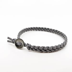 May include: A dark gray braided bracelet featuring a suede-like texture. The bracelet is secured with a single, circular, dark metallic button closure that holds the braided cord in place, creating a simple and functional loop design.