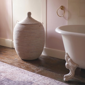 May include: A white wicker laundry basket with a lid and two handles. The basket is sitting on a wooden floor in a bathroom with a white clawfoot bathtub in the background.