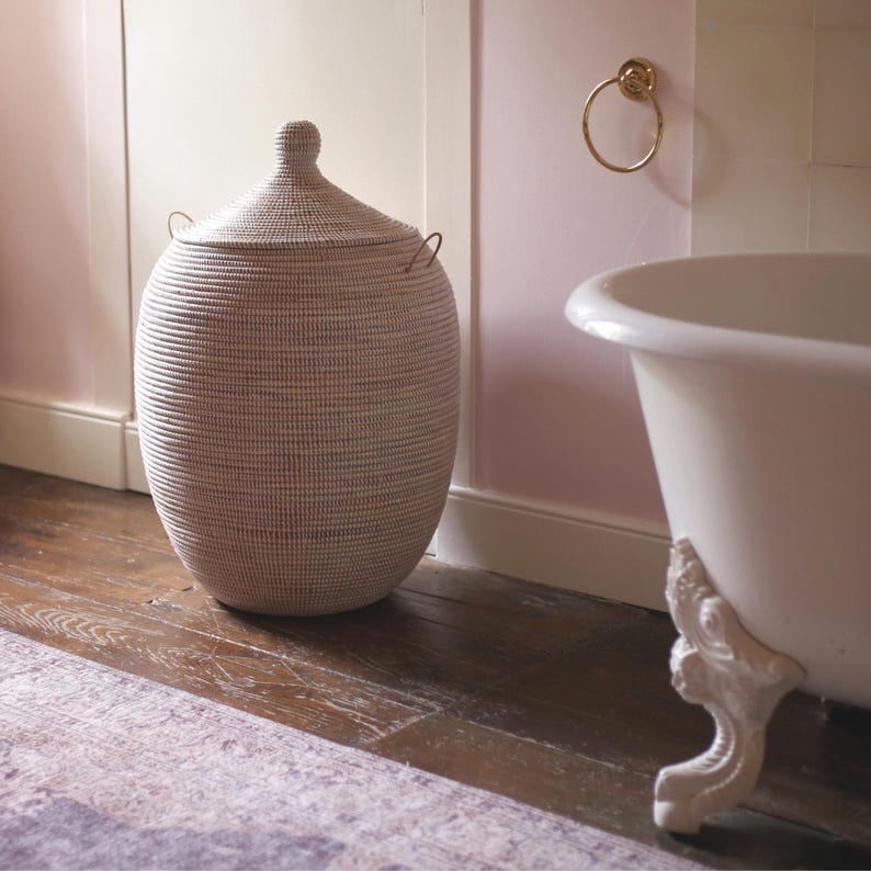 May include: A white wicker laundry basket with a lid and two handles. The basket is sitting on a wooden floor in a bathroom with a white clawfoot bathtub in the background.