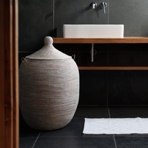 May include: A white woven laundry basket with a lid and gold handles. The basket is in a bathroom with a white sink and a white bath mat.