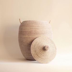 May include: A large, round, woven basket with a lid. The basket is made of natural materials and has a light brown colour. The lid is also woven and has a small knob on top.