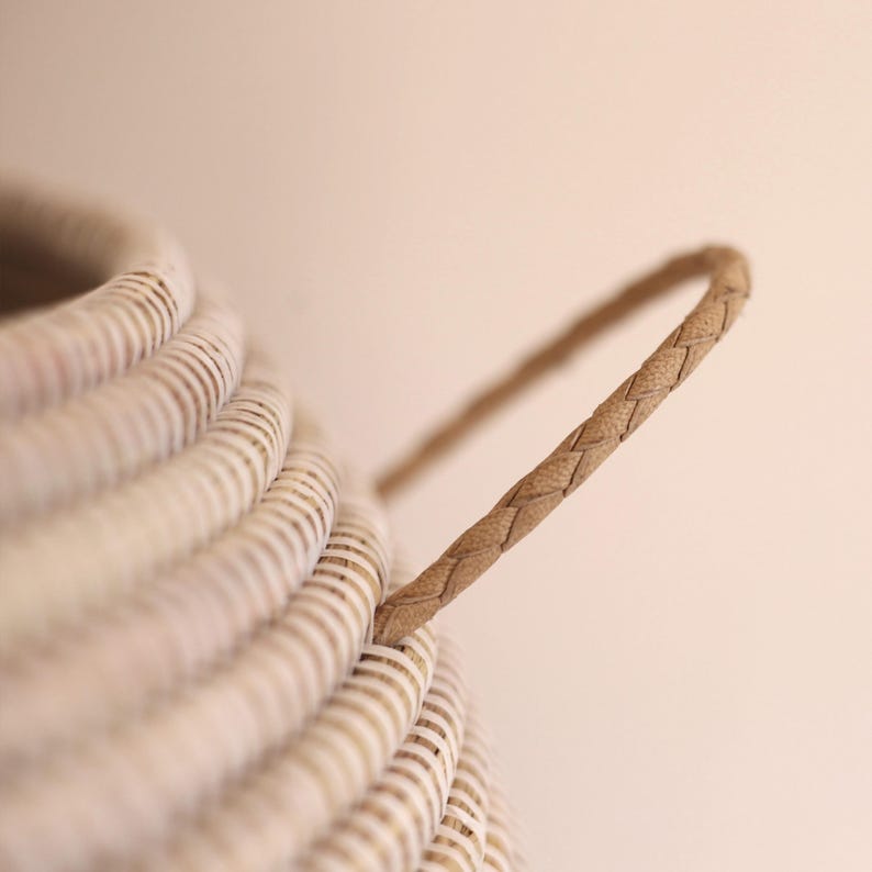 May include: A white wicker basket with a woven leather handle. The basket is made from natural materials and has a rustic look.