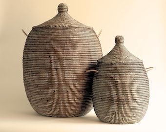 May include: Two woven storage baskets with lids and leather handles. The larger basket is brown and the smaller basket is grey.