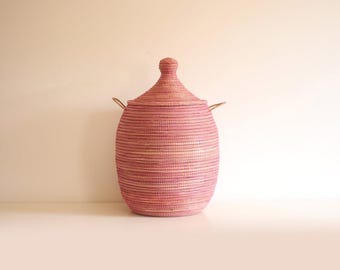 May include: A tall, cylindrical woven basket in a soft pink hue, featuring a matching conical lid with a small knob handle at the top. The basket is constructed from coiled material with a visible horizontal texture and two small side handles.