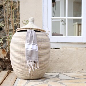 May include: A large, white, woven storage basket with a lid and handles. The basket is made of natural materials and has a round shape. A grey towel is hanging from the basket.