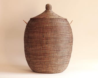 May include: A tall, oval-shaped dark brown woven basket with a matching lid featuring a small knob handle at the top. The basket has a tight, horizontal coil pattern throughout its body and two small, light-coloured protruding side handles.