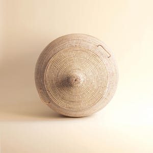 May include: A large, round, white wicker basket with two handles. The basket is woven in a tight pattern and has a slightly domed lid.