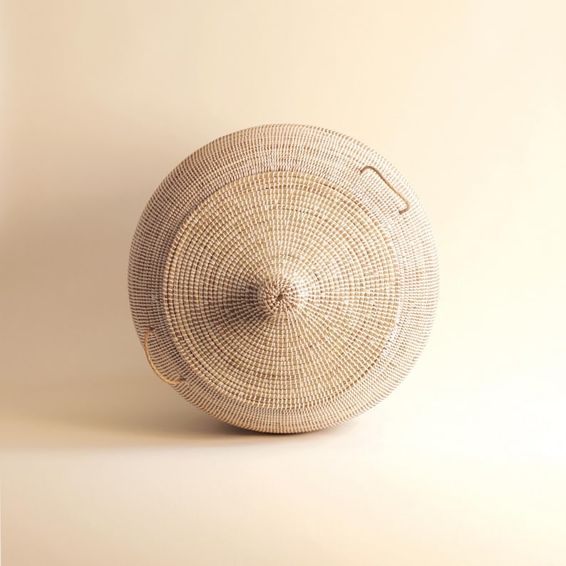 May include: A large, round, white wicker basket with two handles. The basket is woven in a tight pattern and has a slightly domed lid.