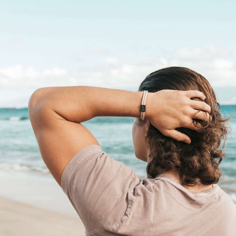 May include: A person wearing a light brown t-shirt with their arm raised, looking out at the ocean. They are wearing a beige and black bracelet on their wrist.
