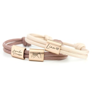 Könnte beinhalten: Zwei verstellbare Kordelarmbänder mit metallischen rechteckigen Perlen. Ein Armband hat eine beige Kordel mit einer Perle, die den Namen Lina und ein Herz trägt. Das andere hat eine braune Kordel mit Perlen, die die Namen Jennifer und Vanessa sowie ein Versprechen zeigen.