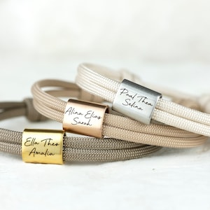 Könnte beinhalten: Drei personalisierte Armbänder mit verschiedenen Namen, die auf jedes Armband graviert sind. Die Armbänder bestehen aus beigefarbenem und weißem Kordel und haben eine goldene, roségoldene und silberne Metallperle.