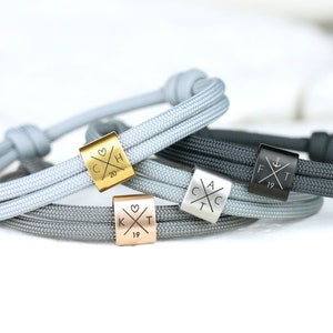 Könnte beinhalten: Drei personalisierte Armbänder mit verschiedenen farbigen Schnüren und Metallanhängern. Die Anhänger sind mit Initialen und einem Herzen, einem Anker und einem Kreuz graviert. Die Armbänder sind grau, schwarz und silberfarben.