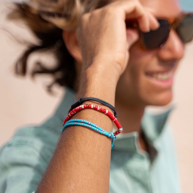 May include: A set of three bracelets worn on the wrist. The top bracelet is a thin black cord. The middle bracelet features red and white cylindrical beads. The bottom bracelet is a bright blue cord with a white speckled pattern and a knot.