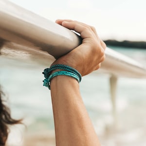 Könnte beinhalten: Ein Arm, der einen Stapel türkisfarbener und schwarzer Seil-Armbänder trägt. Die Person hält ein Surfbrett über ihrem Kopf.