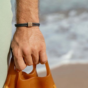 Könnte beinhalten: Eine Person trägt ein schwarz-graues Armband mit einem Kompass-Design. Das Armband besteht aus zwei Schnüren. Die Person hält ein Paar braune Gummiflossen.