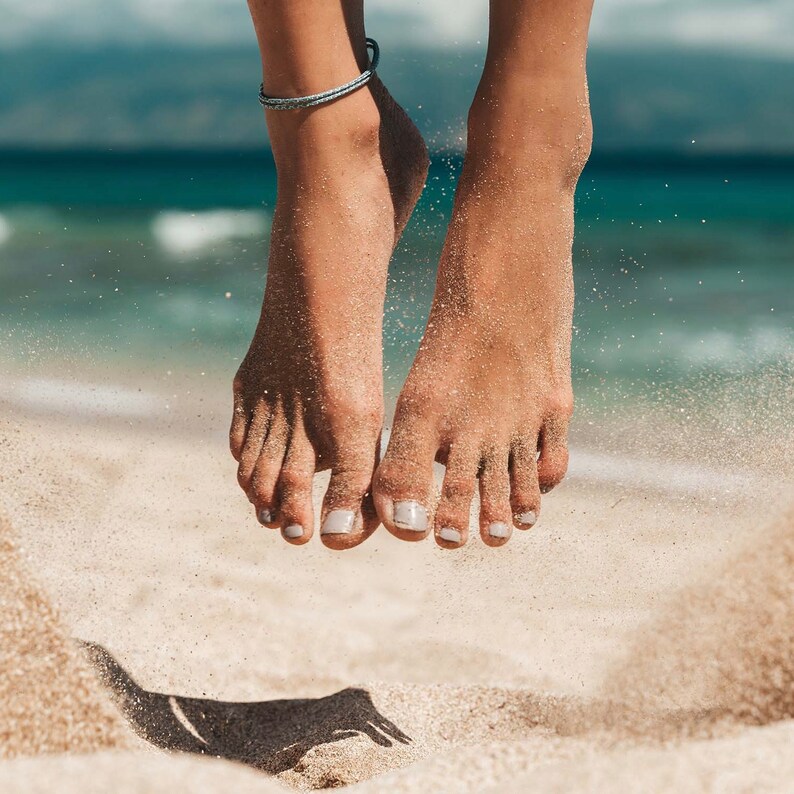 May include: A thin, braided blue and white cord anklet worn around the ankle. The feet are covered in fine sand, with light-colored nail polish on the toes, captured in a mid-air position above a sandy beach with ocean waves in the background.