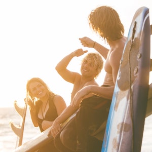 May include: A blue and white surfboard with three fins stands upright next to a group of three people. The surfboard features a smooth, curved surface and a pointed nose, positioned against a bright, sunlit coastal backdrop at golden hour.