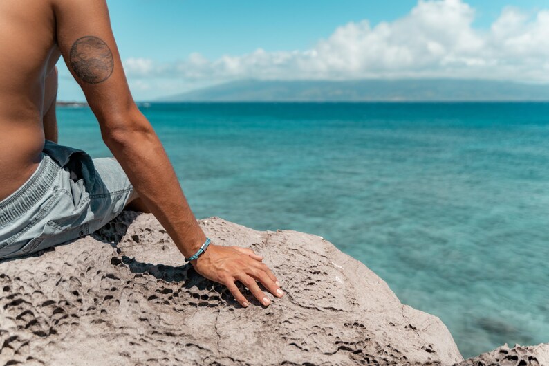 May include: A person sits on a rocky outcropping overlooking a blue ocean. The person is wearing a blue and gray pair of shorts and has a blue bracelet on their wrist.