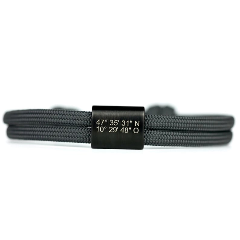 May include: A black double-strand bracelet with a black metal tag engraved with the coordinates "47&deg; 35' 31" N 10&deg; 29' 48" O".