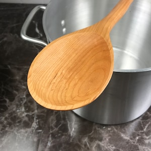 May include: A wooden spoon with a natural finish, resting inside a silver pot. The spoon is made of light-colored wood and has a smooth, rounded surface.