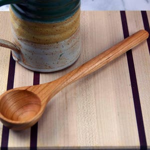 May include: A wooden spoon with a scoop on one end, resting on a wooden cutting board with purple stripes. The spoon is made of light brown wood and has a natural finish.