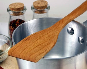 May include: A wooden spatula resting in a silver saucepan. The spatula is made of light brown wood and has a rounded shape. The saucepan is made of aluminum and has a shiny surface.