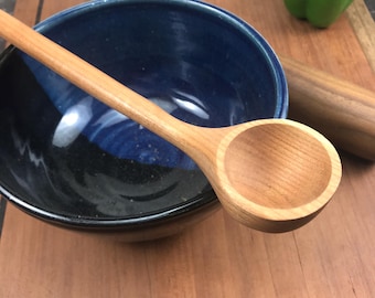May include: A wooden spoon with a rounded bowl, resting in a blue and black ceramic bowl. The spoon is made of light-colored wood and has a smooth, polished finish.