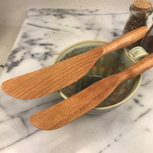 May include: Two wooden butter knives in a ceramic bowl. The knives are made of light brown wood and have a smooth, rounded handle. The bowl is a light blue color with a green and red glaze.