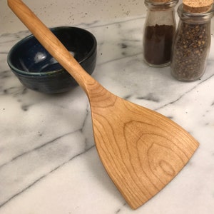 May include: A wooden spatula with a light brown finish and a wide, flat blade. The spatula is made of cherry wood and has a natural grain pattern.