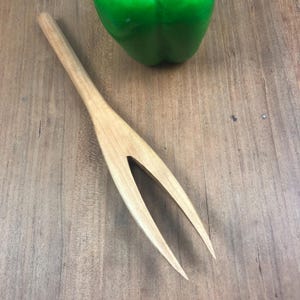 May include: A wooden salad fork with two prongs. The fork is light brown and has a smooth finish. It is lying on a wooden surface.