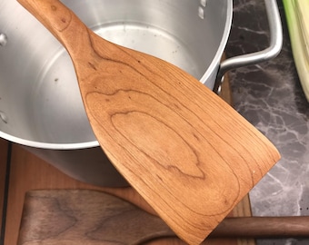 May include: Two wooden kitchen spatulas, one is cherry wood and the other is walnut wood. The cherry wood spatula is resting on the edge of a silver pot.