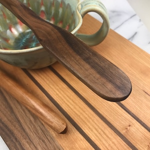 May include: Two wooden kitchen utensils, a spatula and a spoon, are lying on a wooden cutting board with dark brown stripes. The spatula is made of dark brown wood and has a rounded end. The spoon is made of light brown wood and has a rounded end.