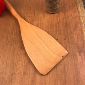 May include: A wooden spatula with a light brown color and a slightly rounded shape. The spatula is lying on a wooden surface.