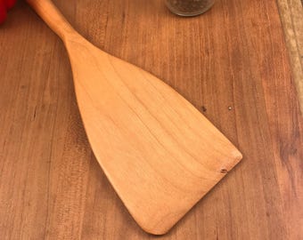May include: A wooden spatula with a light brown color and a slightly rounded shape. The spatula is lying on a wooden surface.