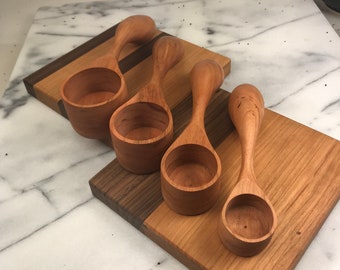 May include: Four wooden measuring spoons with handles, made of cherry wood, on a wooden cutting board with a walnut inlay. The spoons are arranged in a row, with the largest spoon on the left and the smallest spoon on the right.