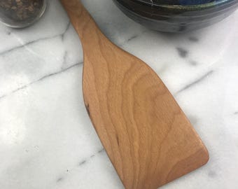 May include: A wooden spatula with a light brown color and a smooth surface. The spatula is made of cherry wood and has a slightly curved shape.