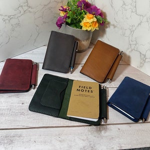 May include: A collection of leather notebooks in various colors, including burgundy, dark brown, and navy blue. Each notebook has a pen holder and a pen. One notebook is open, revealing a "Field Notes" booklet.