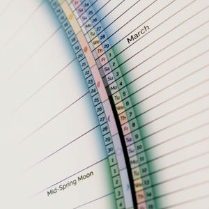 May include: A close-up of a calendar page for March, with a colorful moon phase tracker on the left side. The tracker shows the moon phases for the month, with a crescent moon, full moon, and new moon symbols. The calendar is divided into days of the week, with the days of the week labeled in English. The calendar is printed on white paper.