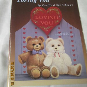 May include: A book cover titled "Loving You" by Camille & Sue Scheewe. The cover features two teddy bears, one brown with a blue bow tie and the other white. A red heart with the words "Loving You" is above the bears.
