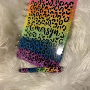 May include: A glittery notebook and pen set with a rainbow leopard print design. The notebook and pen have the name "Emersyn" written on them. The notebook is spiral-bound.
